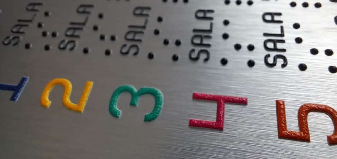 Detalle de mapa háptico: números en relieve de colores y puntos Braille sobre una superficie metálica cepillada.