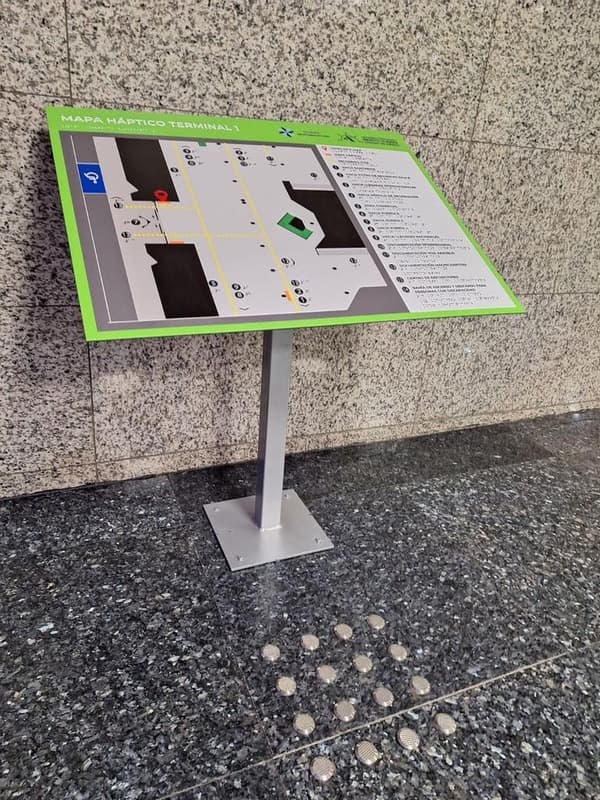 Mapa háptico con marco verde montado sobre un pedestal metálico de piso, frente a un muro de granito.